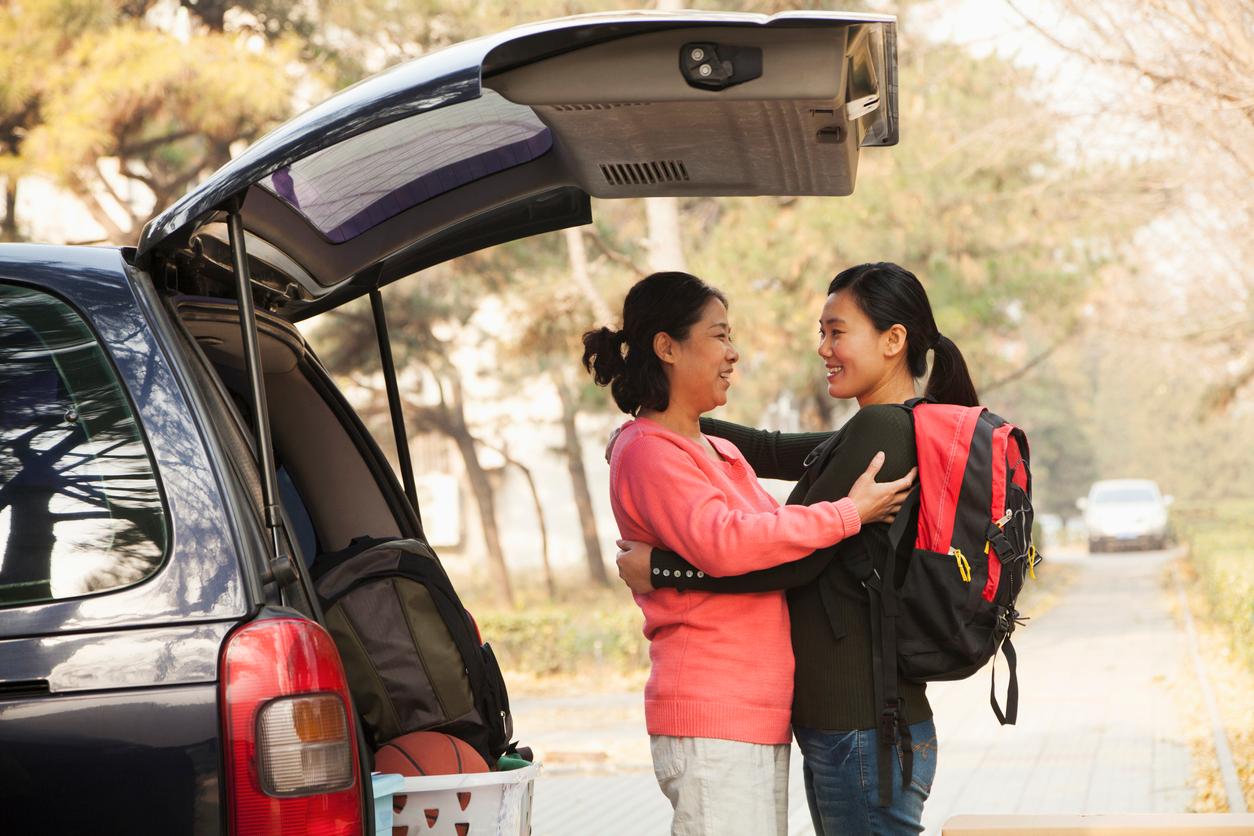 An Asian mother and daughter embrace beside a car trunk loaded with college move-in supplies. The mother wears a coral pink sweater while the daughter carries a backpack. Golden afternoon light captures this bittersweet farewell moment.