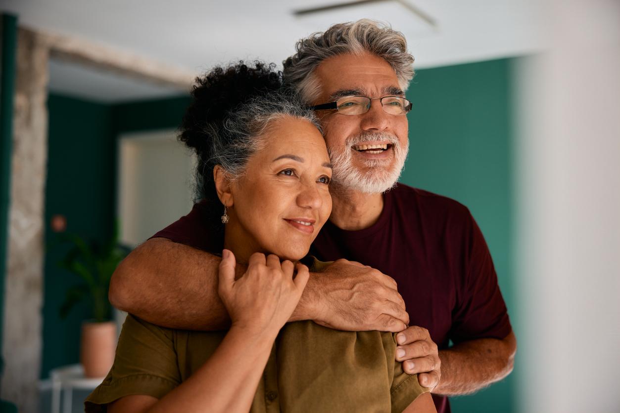 A loving couple in their 50s-60s shares an affectionate embrace in their modern home. The gray-haired man wraps his arms around the woman from behind, both laughing joyfully. A teal accent wall and natural lighting convey comfort and partnership.