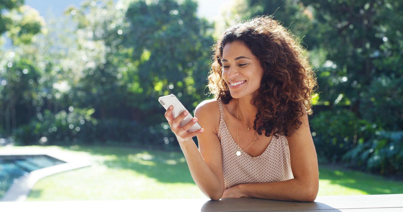 A woman with long, curly brown hair smiles warmly while looking at her smartphone on an outdoor patio. She wears a beige polka dot tank top and gold necklace. A sunlit backyard with lawn, trees, and pool creates a relaxed summer atmosphere.
